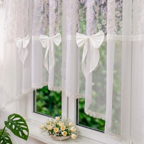 Biała firana woalowa Estella zawieszona w oknie, pokazująca delikatny kwiatowy haft oraz dekoracyjne upięcie satynową kokardą.